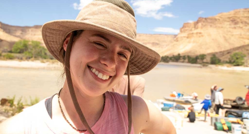 A person wearing a sun hat smiles in front of a river landscape with canyon walls in the background. 